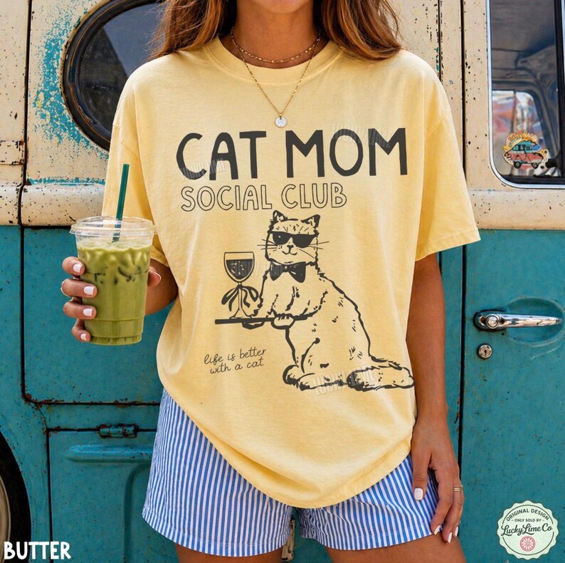 K&ouml;nnte beinhalten: Ein blassgelbes Kurzarm-T-Shirt mit schwarzem Aufdruck CAT MOM SOCIAL CLUB und einer Grafik einer Katze mit Sonnenbrille und Fliege, die ein Weinglas h&auml;lt, sowie dem darunter gedruckten Text Life is better with a cat.