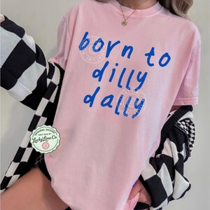 May include: A light pink t-shirt with the phrase "born to dilly dally" in blue script. The shirt has a crew neck and short sleeves. A black and white checkered cardigan is draped over the shoulders. A Lucky Lime Co. logo is visible.
