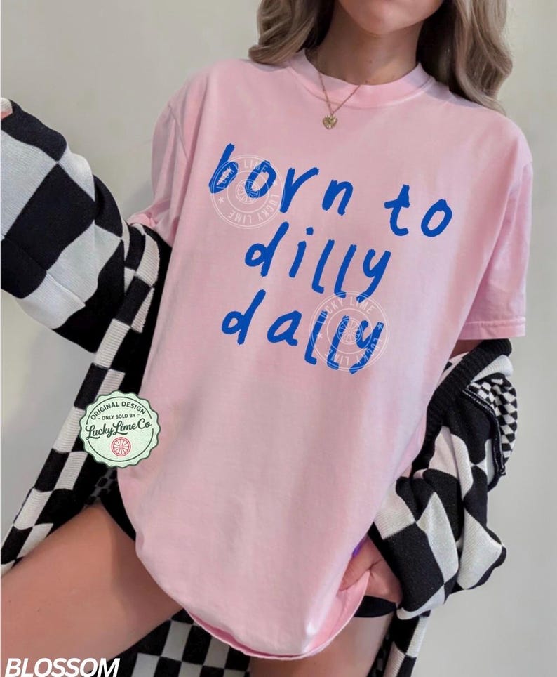 May include: A light pink t-shirt with the phrase "born to dilly dally" in blue script. The shirt has a crew neck and short sleeves. A black and white checkered cardigan is draped over the shoulders. A Lucky Lime Co. logo is visible.