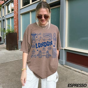 May include: A brown t-shirt with a blue graphic print of London landmarks, including a double-decker bus, a phone booth, a crown, and the words "LONDON".