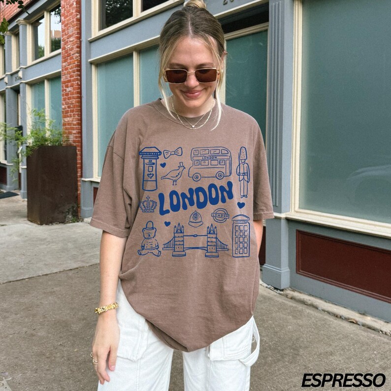 May include: A brown t-shirt with a blue graphic print of London landmarks, including a double-decker bus, a phone booth, a crown, and the words "LONDON".