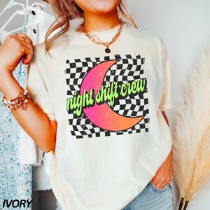 May include: A white t-shirt with a black and white checkered design and a pink crescent moon. The text "night shift crew" is written in green on the moon. The shirt is a light ivory color.