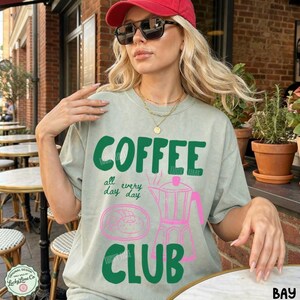 Peut inclure: Un t-shirt &agrave; col rond vert sauge avec le texte COFFEE CLUB en lettres vertes grasses. Sous le texte se trouve un dessin au trait rose d'une cafeti&egrave;re italienne et d'une p&acirc;tisserie, avec les mots all day every day en petites lettres vertes.