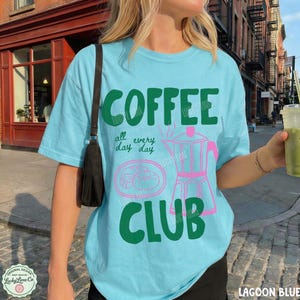 Peut inclure: Un t-shirt bleu clair avec le texte vert fonc&eacute; COFFEE CLUB et la phrase all every day day. Le motif comprend un contour rose d'une cafeti&egrave;re italienne et d'une p&acirc;tisserie. Une bandouli&egrave;re de sac noire est visible sur l'&eacute;paule gauche.