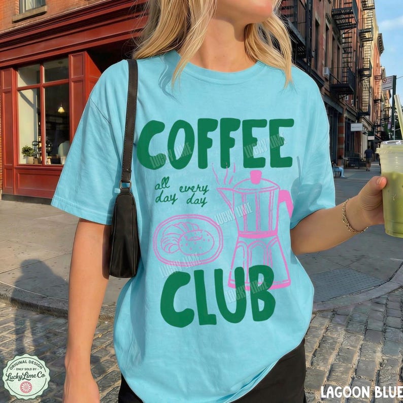 Peut inclure: Un t-shirt bleu clair avec le texte vert fonc&eacute; COFFEE CLUB et la phrase all every day day. Le motif comprend un contour rose d'une cafeti&egrave;re italienne et d'une p&acirc;tisserie. Une bandouli&egrave;re de sac noire est visible sur l'&eacute;paule gauche.