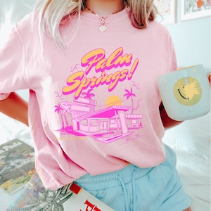 May include: Pink t-shirt with a retro graphic design featuring the text "Palm Springs!" and a stylised illustration of a building with palm trees and a sunset.
