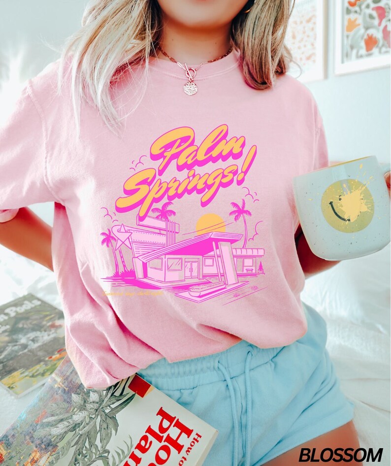 May include: Pink t-shirt with a retro graphic design featuring the text "Palm Springs!" and a stylised illustration of a building with palm trees and a sunset.