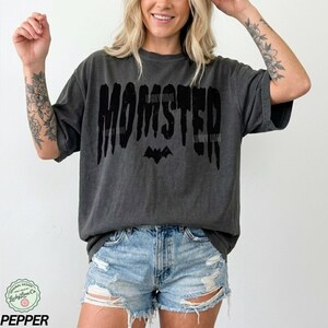 May include: Dark gray t-shirt with the word "MOMSTER" in a dripping font, a bat graphic, and the text "LUCKY LIME". The shirt is worn with denim shorts. The shirt is from Lucky Lime Co.