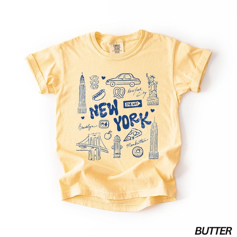 May include: Butter yellow t-shirt with a blue graphic of New York City landmarks, including the Statue of Liberty, the Empire State Building, and the Brooklyn Bridge. The words "New York" are prominently displayed.