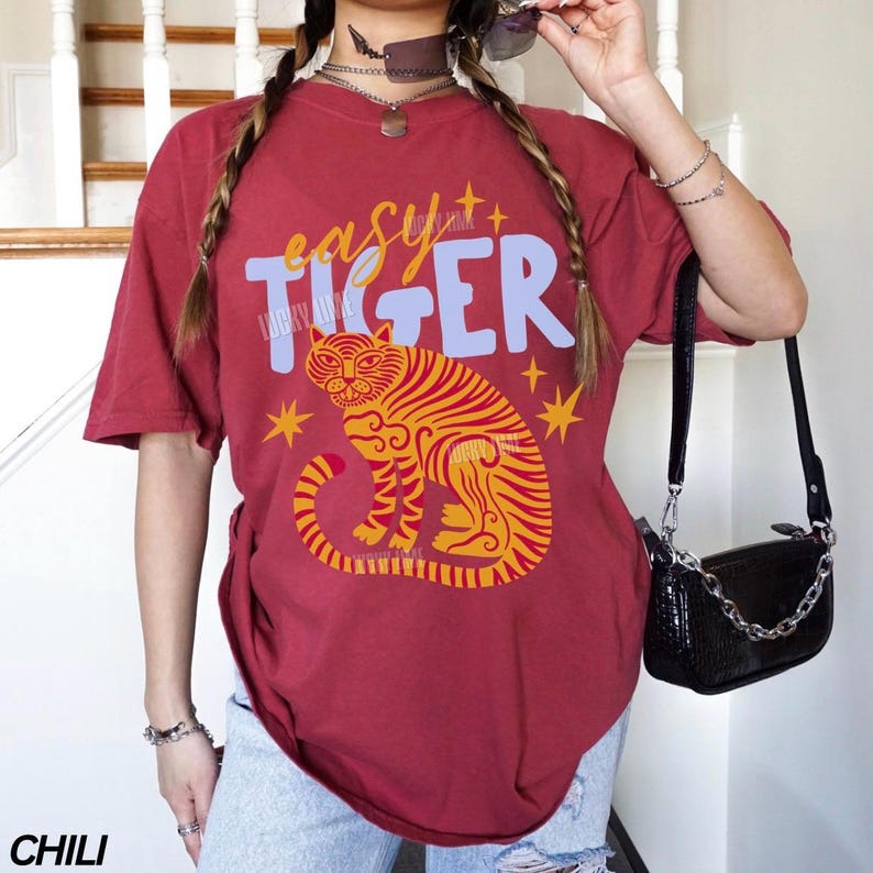 May include: Chili-colored t-shirt with the text "easy tiger" in blue and yellow. The shirt features a tiger illustration in yellow and orange. The word "CHILI" is printed at the bottom left corner of the shirt.