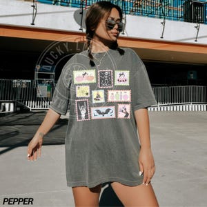 May include: Dark gray t-shirt featuring a collection of Halloween-themed postage stamp designs. The stamps showcase pumpkins, skeletons, spiders, witch hats, potion bottles, bats, and more. The word "PEPPER" is printed at the bottom.