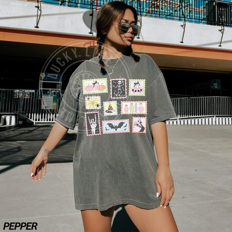May include: Dark gray t-shirt featuring a collection of Halloween-themed postage stamp designs. The stamps showcase pumpkins, skeletons, spiders, witch hats, potion bottles, bats, and more. The word "PEPPER" is printed at the bottom.