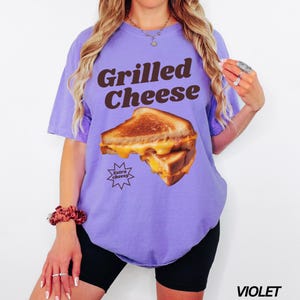 May include: A purple t-shirt with the text "Grilled Cheese" in brown letters. The shirt features a graphic of a grilled cheese sandwich with the text "Extra Cheesy" in a star shape.