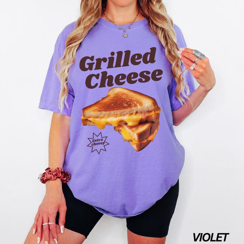 May include: A purple t-shirt with the text "Grilled Cheese" in brown letters. The shirt features a graphic of a grilled cheese sandwich with the text "Extra Cheesy" in a star shape.