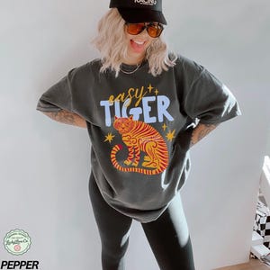 May include: Dark gray t-shirt with the words "easy TIGER" in blue and yellow. The shirt features a tiger illustration in red and yellow. The model is wearing a black cap and sunglasses.