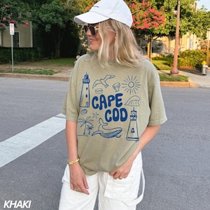Peut inclure: Un t-shirt vert kaki avec un motif graphique bleu repr&eacute;sentant un phare, une baleine, une coquille, une &eacute;toile de mer, une vague et le texte "Cape Cod".