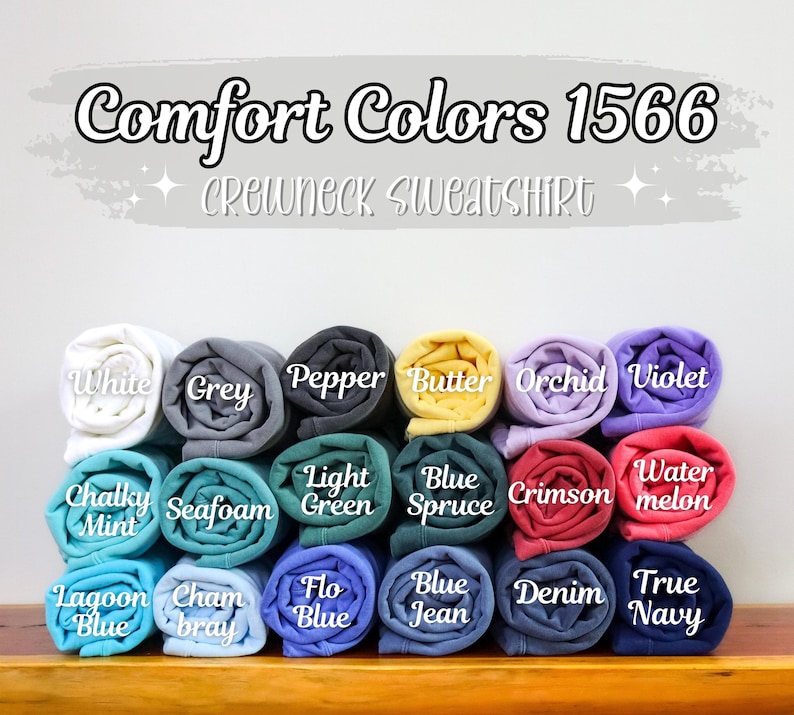 May include: A display of rolled-up Comfort Colors 1566 crewneck sweatshirts in various colors, including white, grey, and blue. The image features the text "Comfort Colors 1566 Crewneck Sweatshirt" at the top, with color names labeled below each rolled shirt.