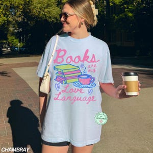 以下が含まれることがあります： ピンクと緑で「Books are my Love Language」と書かれた水色のTシャツ。グラフィックには本の山、ティーカップ、ハートが含まれています。 ゆったりとしたカジュアルなフィット感のシャツです。