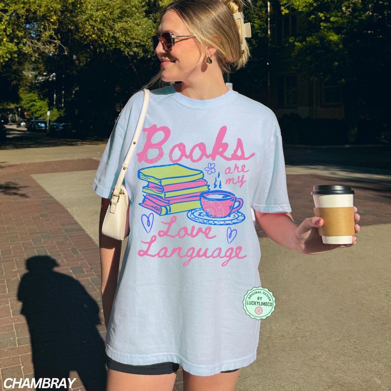 以下が含まれることがあります： ピンクと緑で「Books are my Love Language」と書かれた水色のTシャツ。グラフィックには本の山、ティーカップ、ハートが含まれています。 ゆったりとしたカジュアルなフィット感のシャツです。