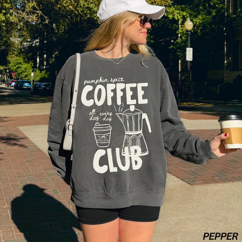 May include: A charcoal gray sweatshirt with the words "COFFEE CLUB" in white, along with a coffee pot and a to-go cup graphic. The sweatshirt also has the words "pumpkin spice" and "all every day day". The person is wearing a white cap and sunglasses.