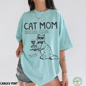 K&ouml;nnte beinhalten: Ein helles mintgr&uuml;nes T-Shirt mit schwarzem Text CAT MOM SOCIAL CLUB und Life is better with a cat. Die Grafik zeigt eine Katze mit Sonnenbrille und Fliege, die ein Weinglas auf einem Tablett h&auml;lt. Das T-Shirt hat kurze &Auml;rmel.