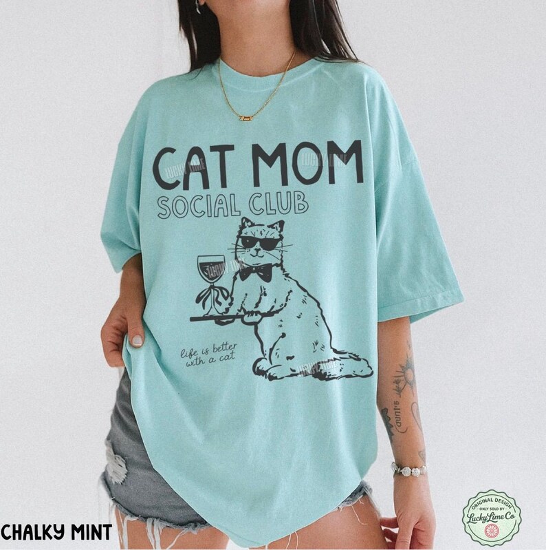 K&ouml;nnte beinhalten: Ein helles mintgr&uuml;nes T-Shirt mit schwarzem Text CAT MOM SOCIAL CLUB und Life is better with a cat. Die Grafik zeigt eine Katze mit Sonnenbrille und Fliege, die ein Weinglas auf einem Tablett h&auml;lt. Das T-Shirt hat kurze &Auml;rmel.