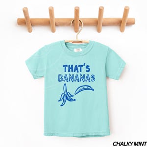 Op de afbeelding: Een lichtblauw t-shirt met een blauwe afbeelding van een banaan en de tekst "THAT'S BANANAS". Het shirt hangt aan een houten rek tegen een witte achtergrond. De kleur van het shirt wordt beschreven als "krijtachtig mint".