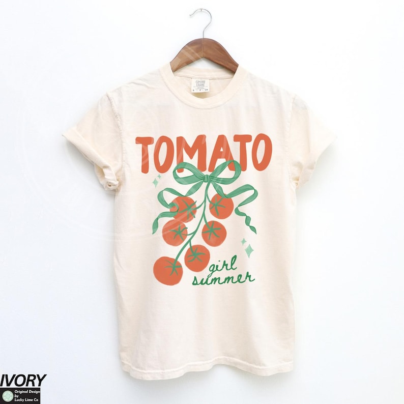 May include: Ivory-colored t-shirt with the word "TOMATO" in orange block letters. Below, a green bow and ribbon design with a cluster of orange tomatoes and the words "girl summer" in green.