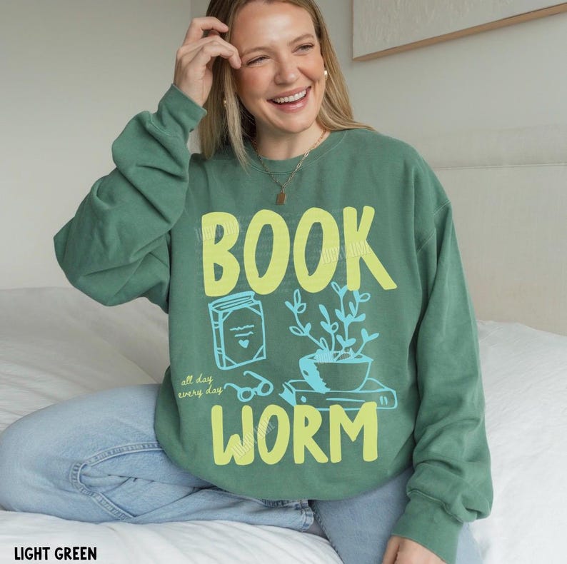 May include: Light green sweatshirt with the words "BOOK WORM" in yellow, along with a book, glasses, and a potted plant graphic in blue. The text "all day every day" is also included. The sweatshirt is a comfortable, casual style.