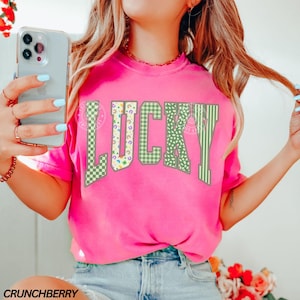 May include: A bright pink t-shirt with the word "LUCKY" in large, green, patterned letters. The letters feature a mix of checkered, floral, and striped designs. The text "CRUNCHBERRY" is printed at the bottom left corner of the shirt.