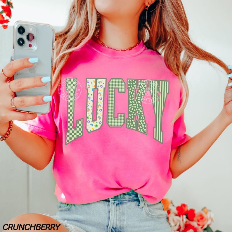 May include: A bright pink t-shirt with the word "LUCKY" in large, green, patterned letters. The letters feature a mix of checkered, floral, and striped designs. The text "CRUNCHBERRY" is printed at the bottom left corner of the shirt.