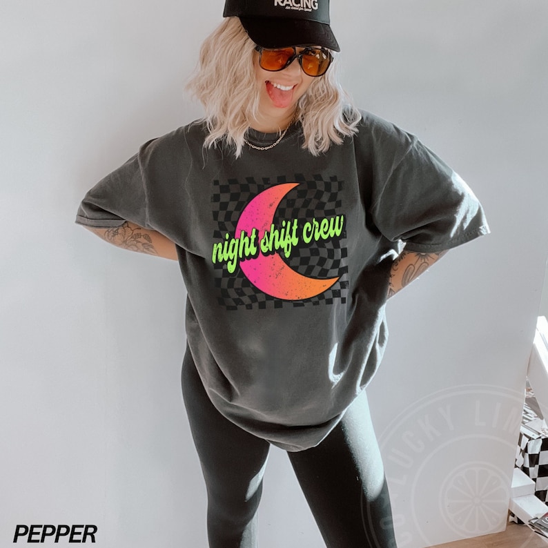 May include: A black t-shirt with a graphic of a crescent moon with a pink and orange gradient on a black and white checkered background. The text "night shift crew" is written in green on the moon.
