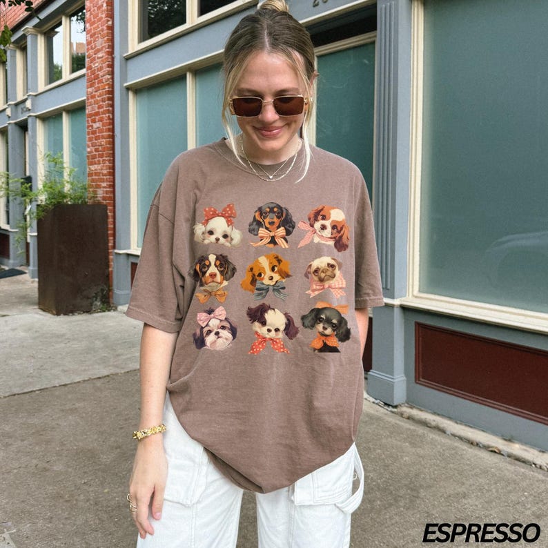 May include: A brown t-shirt with a graphic of various dogs wearing bows. The dogs are all different breeds and have different expressions. The shirt is being worn by a person with long brown hair and glasses.
