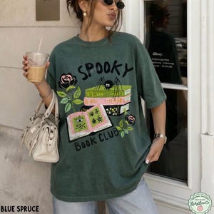 Op de afbeelding: Blauwspar T-shirt met een grafisch ontwerp met de tekst "Spooky Book Club", een spin, boeken en rozen. Het shirt heeft een relaxte pasvorm en korte mouwen.