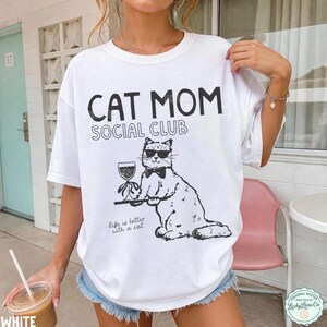 K&ouml;nnte beinhalten: Ein wei&szlig;es Kurzarm-T-Shirt mit schwarzem Aufdruck CAT MOM SOCIAL CLUB. Darunter befindet sich eine Grafik einer Katze mit Sonnenbrille und Fliege, die ein Weinglas auf einem Tablett h&auml;lt, sowie der Text Life is better with a cat.