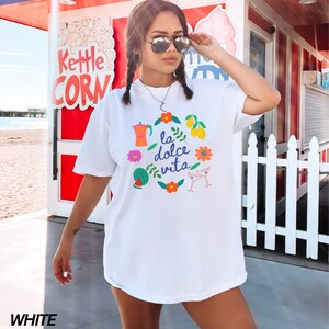 Pu&ograve; includere: Maglietta bianca con un design grafico colorato che presenta la frase "la dolce vita" circondata da limoni, fiori, una fetta di anguria, un bicchiere da cocktail e una caffettiera.