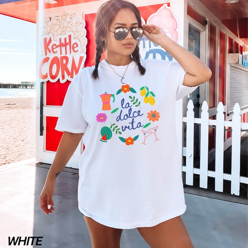 Pu&ograve; includere: Maglietta bianca con un design grafico colorato che presenta la frase "la dolce vita" circondata da limoni, fiori, una fetta di anguria, un bicchiere da cocktail e una caffettiera.