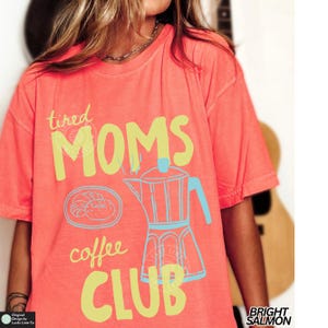 May include: Bright salmon-colored t-shirt with the words "tired MOMS coffee CLUB" in yellow and blue. The shirt features a coffee pot and a croissant graphic. The shirt is a relaxed fit.