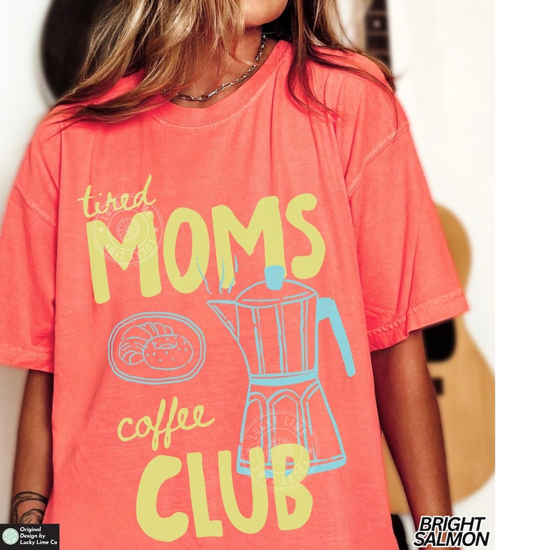 May include: Bright salmon-colored t-shirt with the words "tired MOMS coffee CLUB" in yellow and blue. The shirt features a coffee pot and a croissant graphic. The shirt is a relaxed fit.