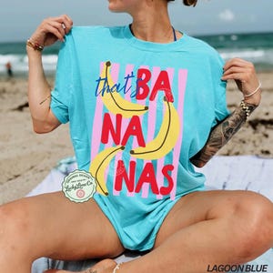Puede incluir: Camiseta azul laguna con la frase "that's BANANAS" en un dise&ntilde;o divertido. El dise&ntilde;o presenta pl&aacute;tanos amarillos y rayas rosas. Una prenda informal para la playa o las vacaciones.