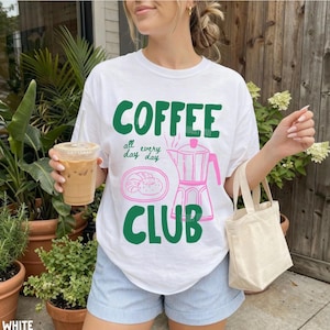 Peut inclure: Un t-shirt blanc &agrave; manches courtes avec le texte vert COFFEE CLUB, les mots all day every day, un dessin au trait rose d'un croissant sur une assiette et un dessin au trait rose d'une cafeti&egrave;re moka sur le devant du v&ecirc;tement.