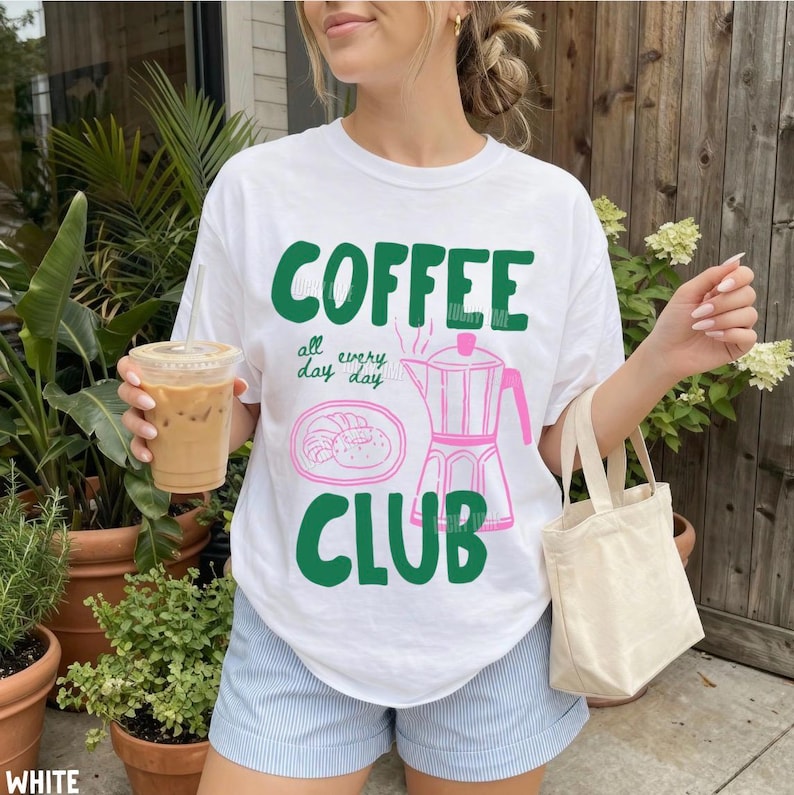Peut inclure: Un t-shirt blanc &agrave; manches courtes avec le texte vert COFFEE CLUB, les mots all day every day, un dessin au trait rose d'un croissant sur une assiette et un dessin au trait rose d'une cafeti&egrave;re moka sur le devant du v&ecirc;tement.