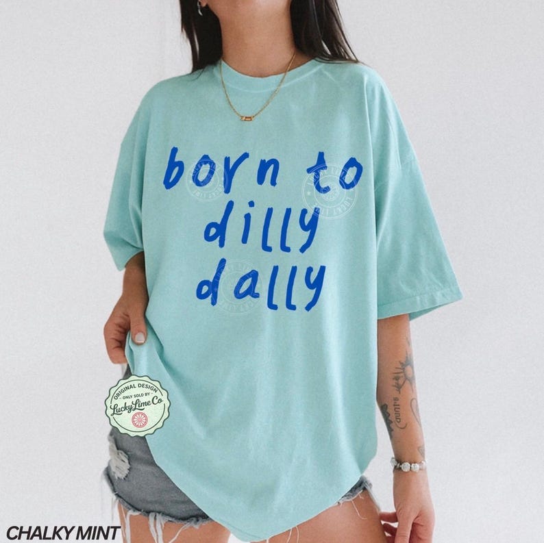 May include: A light blue t-shirt with the phrase "born to dilly dally" in blue lettering. The shirt has a relaxed fit and short sleeves. The text is in a playful, handwritten style. The shirt is labeled "Chalky Mint".