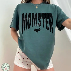 May include: Blue spruce-colored t-shirt with the word "MOMSTER" in black, featuring a bat graphic. The shirt is a relaxed fit. The text "Lucky Lime Co" is also visible.