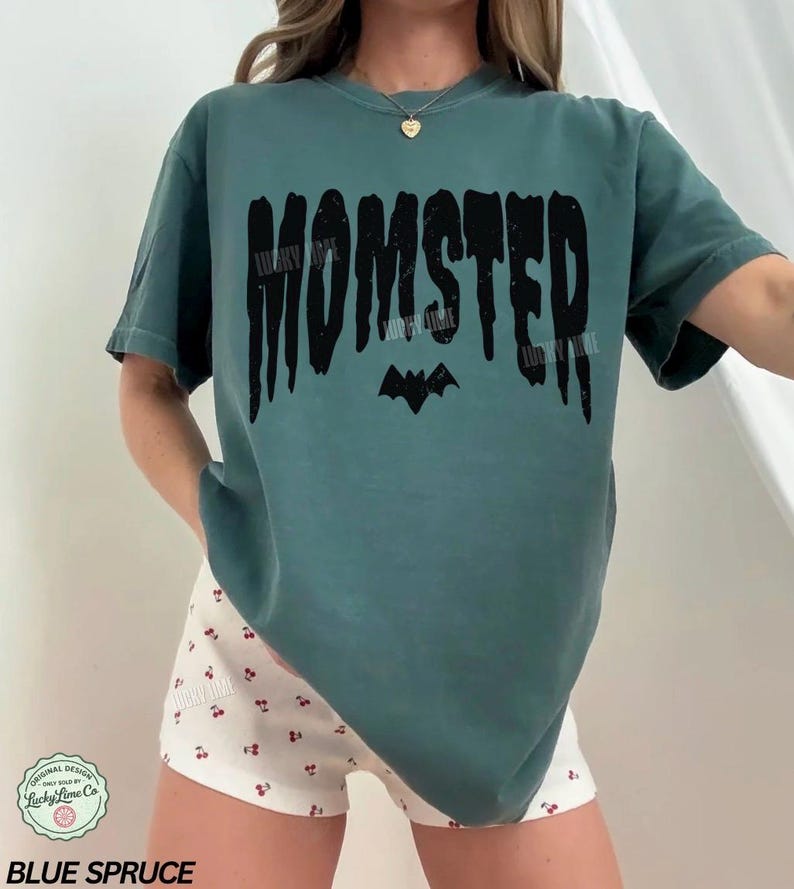May include: Blue spruce-colored t-shirt with the word "MOMSTER" in black, featuring a bat graphic. The shirt is a relaxed fit. The text "Lucky Lime Co" is also visible.