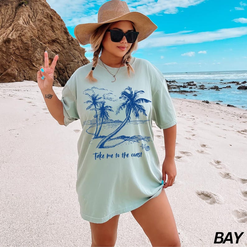 May include: A light blue t-shirt with a graphic of palm trees and the text "Take me to the coast". The model is wearing a straw hat and sunglasses and is standing on a beach.