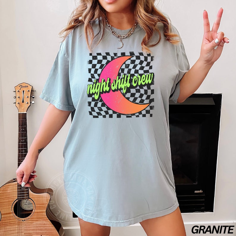 May include: A light blue t-shirt with a graphic of a crescent moon on a black and white checkered background. The text "night shift crew" is written in green and pink letters on the moon.