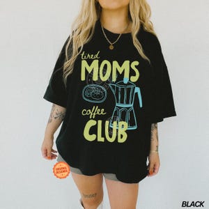 May include: Black t-shirt with a graphic design featuring the words "tired MOMS coffee CLUB" in green and yellow. The design includes a coffee pot and a pastry. The shirt is a casual, oversized fit.