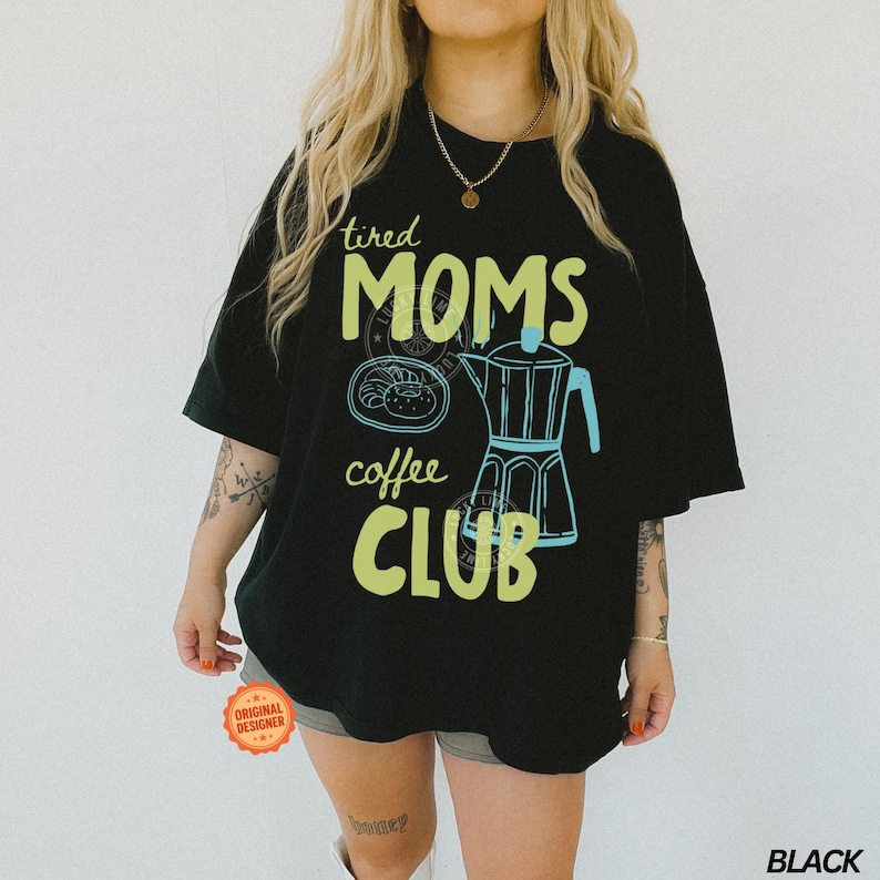 May include: Black t-shirt with a graphic design featuring the words "tired MOMS coffee CLUB" in green and yellow. The design includes a coffee pot and a pastry. The shirt is a casual, oversized fit.