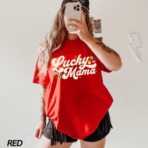 Peut inclure: T-shirt rouge avec le texte "Lucky Mama" dans une police r&eacute;tro avec des tr&egrave;fles et des c&oelig;urs color&eacute;s.